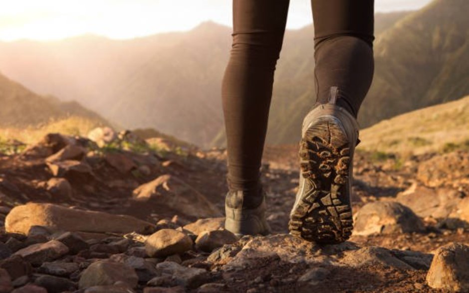A pair of sneaker-clad feet walking away from the camera on a rough-terrain trail. There are mountains in the distance.