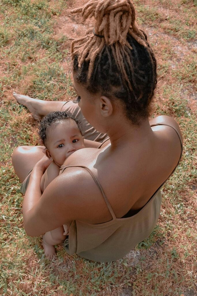 A black woman wearing an olive green tank top and dreadlocks pulled up into a bun, breastfeeding her infant child while sitting on the grass outside. Photograph is taken from over her shoulder, so her face is unseen, but her baby faces the camera directly.