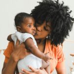 A black woman with medium-length natural hair wearing an orange shirt, holding a newborn baby and giving the baby a kiss on the cheek