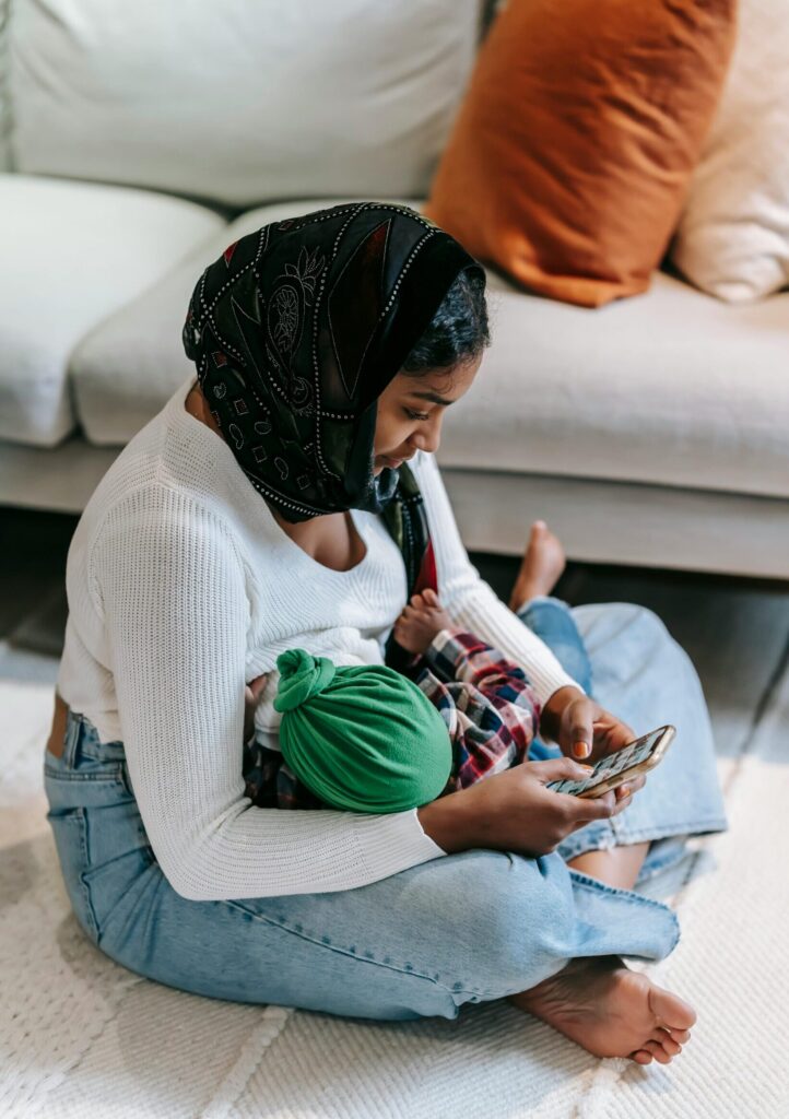 A dark-skinned woman sitting on the floor in front of a couch wearing a white long-sleeved shirt, jeans, and a headscarf. She breastfeeding her baby, who is wearing a green headscarf, and holding a cell phone in her hands.