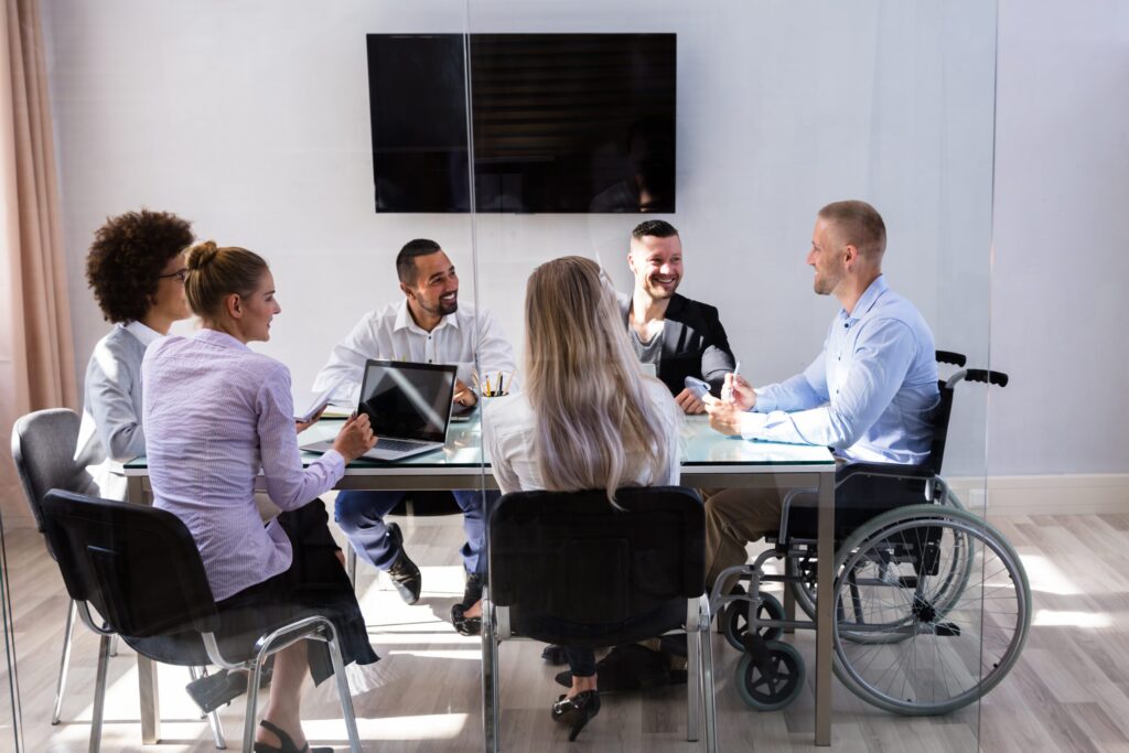 A group of six people sitting in a conference room around a table conversing. They have laptops and notebooks in front of them as though they are working. One is in a wheelchair.