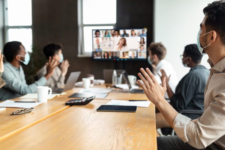 A group of professionals sitting at a conference table with laptops, tablets, calculators, pens, and papers in front of them. At the far end of the conference table there is a screen on the wall where a video call is taking place showing a large number of participants. Those in the room are wearing protective medical masks and waving to the screen.